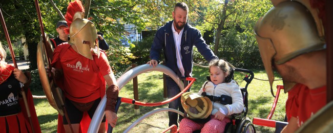 Dziecko na wózku inwalidzkim na podjeździe, obok stoi mężczyźni, w tym kliku w historycznych zbrojach Spartan.