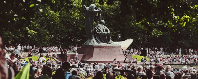 Ludzie siedzią na trawie wokół Pomnika Fryderyka Chopina.