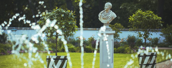 Fragment tryskającej wodą fontanny, w tle egzotyczne drzewka w donicach i marmurowe popiersie, ustawione na cokole.