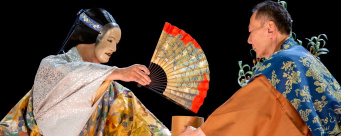 Japoński aktor w masce na twarzy, z wachlarzem w ręku podaje aktorowi w kimonie drewnianą miskę.