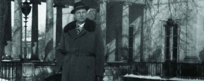 Mężczyzna w płaszczu i kapeluszu stoi przed Pałacem na Wyspie zimą. 