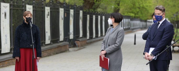 Kobieta w czarnej kurtce, czerwonej spódnicy i maseczce na twarzy stoi przed mikrofonem, na tle plansz w Galerii Plenerowej, z boku obok stoi kobieta i dwóch mężczyzn.