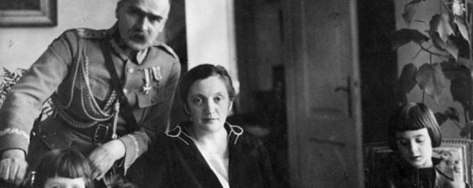 Zdjęcie marszałka Piłsudskiego z rodziną. Marszałek, ubrany w mundur, stoi pośrodku, opierając się lewą ręką o fotel, na którym siedzi jego żona. Po lewej i po prawej stronie siedzą jego dwie córki. 