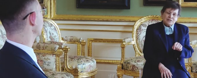 W sali, gdzie na ścianach wiszą obrazy, na ozdobnym fotelu z epoki siedzi starsza kobieta w granatowym kostiumie, naprzeciwko kobiety siedzi odwrócony tyłem mężczyzna. 