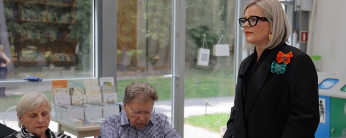 Kobieta w czarnym stroju stoi przy stole, obok, po jej prawej stronie, siedzą przy stole starsza kobieta i mężczyzna.