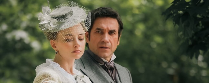 Kobieta w długiej, białej, koronkowej sukni i kapeluszu spaceruje obok mężczyzny w garniturze na tle parku.