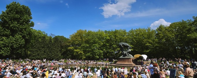 Ludzie siedzią na trawie wokół Pomnika Fryderyka Chopina.