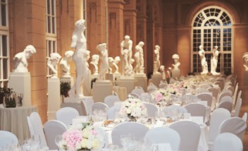 Sala, w której stoją stoły przykryte białym obrusami, a wokół nich krzesła. Pod ścianą stoją rzeźby.
