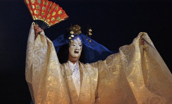 Aktor teatru Nō w kimonie i masce, unoszący ręce, w jednej ręce aktor ma czerwono-złoty wachlarz.
