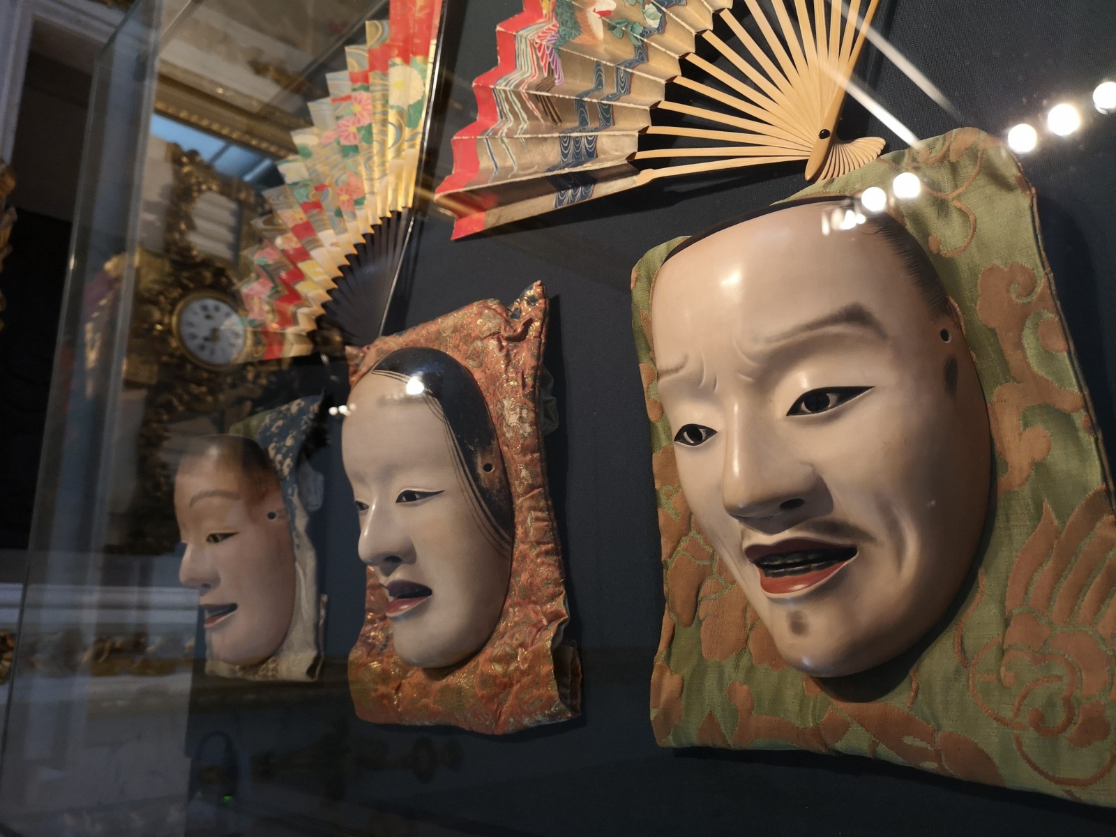 Trzy maski  japońskiego teatru nō wiszące nad czarnej ścianie, nad nim znajdują się ozdobione japońskimi wzorami dwa wachlarze.