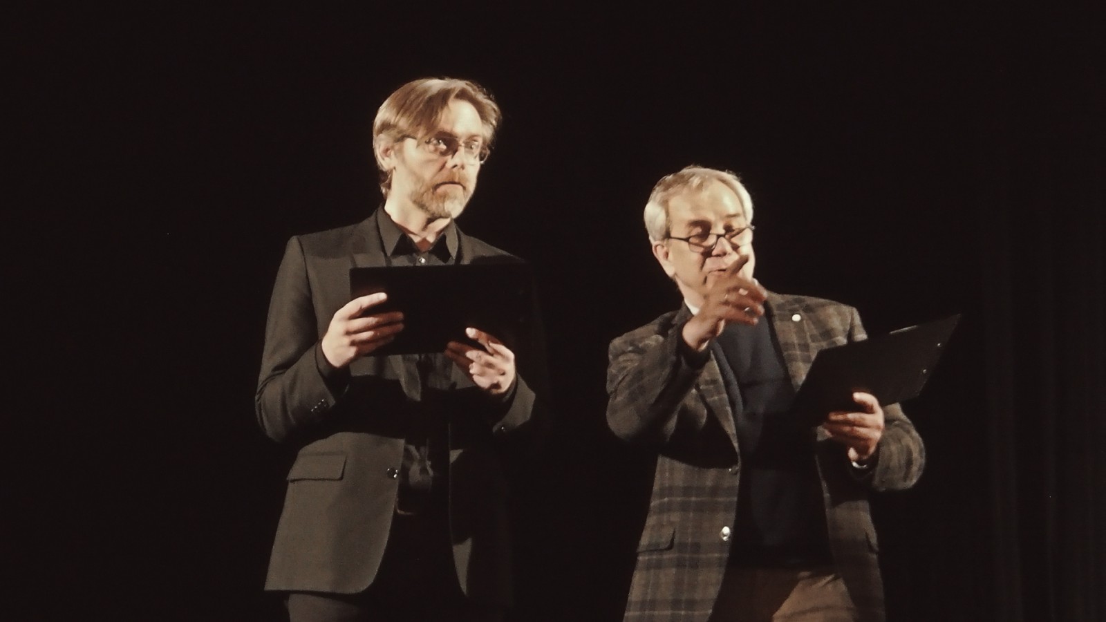 Aktorzy Oskar Hamerski i prof. Jarosław Gajewski stojąc, czytają. Mężczyna z prawej unosi palec wskazujący.