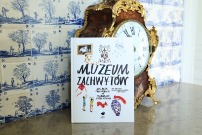 Książka stoi oparta o zegar, na tle ściany ozdobionej niebieskimi malunkami, przedstawijącymi sielski krajobraz.