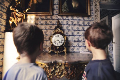 Dwóch chłopców stoi przed ścianą, na której wiszą obrazy. Na komodzie pod ścianą stoi zegar. 