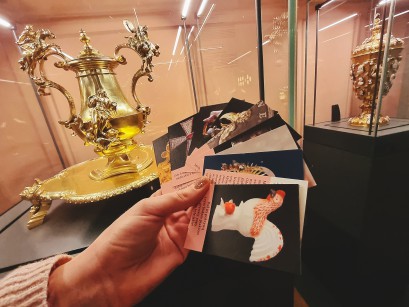 Kobieca ręka trzyma talię kolorowych kart na tle dwóch gablot wystawowych, w których znajdują się bogato zdobione złote wazony. 