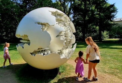 Troje dzieci stoi przy dużej okrągłej białej kuli, która znajduje się w ogrodzie.