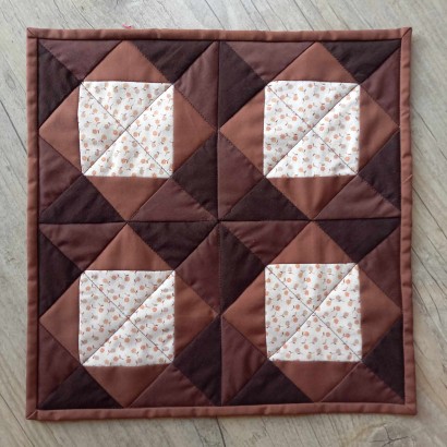 Kawałek brązowej, kwadratowej tkaniny, na której naszyte są cztery małe jasne kwadraty, ułożone w obok siebie w dwóch rzędach. 