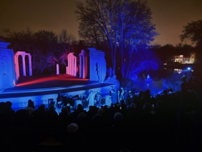 Scena na wolnym powietrzu otoczona kawałkami murów. Jest noc, scena jest podświetlona na niebiesko. Przed sceną widać tłum. 