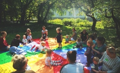 Grupa dzieci i dorosłych siedzi na kolorowym kocu w ogrodzie pod drzewami. 