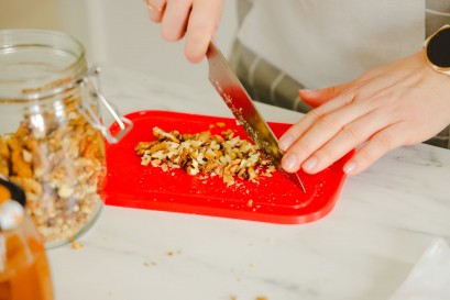 Ręka krojąca nożem orzechy na czerwonej desce. Obok stoi słoik z orzechami.