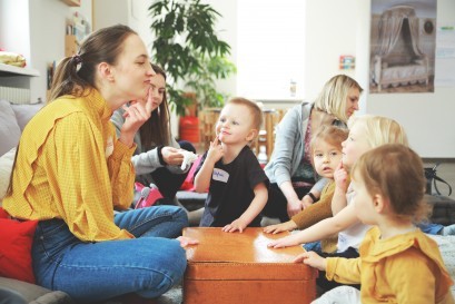 Kobieta siedzi w pomieszczeniu z dziećmi na podłodze. Kobieta dotyka palcem brody, dzieci ją naśladują. Z tyłu za dziećmi siedzą dwie inne kobiety.