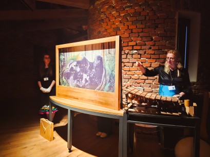 W kwadratowej, drewnianej ramie znajduje się plansza z ilustracją. Po obu stronach ramy stoją kobiety. Kobieta po prawej stronie uderza w drewniany instrument. 