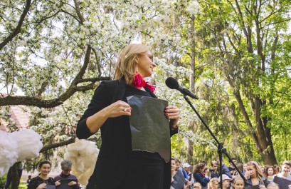 Kobieta z czarną kartką trzymaną w rękach stoi przed mikrofonem. W tle widać tłum ludzi stojący pod drzewami.