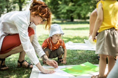 Dziecko i matka w ogrodzie, przykucnęli na trawniku przy rozłożonych na trawie kolorowych planszach. Obok, po prawej, stoi tyłem inne dziecko. 