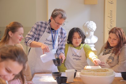 Trzy kobiety, mężczyzna i młody chłopak są zgromadzeni przy stole, na którym stoją pojemniki i gipsowe formy. Mężczyzna nalewa płyn z pojemnika do miseczki. Chłopak sięga ręką po glinianą formę. 