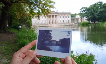 Pałac na Wyspie w otoczeniu drzew i wody. Na tle Pałacu kobiece dłonie trzymają archiwalną fotografię Pałacu na Wyspie.