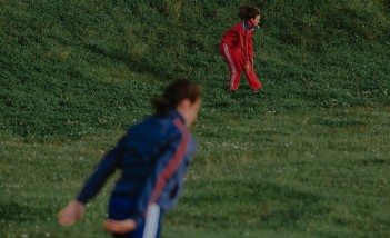 Dwie kobiety w dresach, jedna w niebieskim, druga w czerwonym, tańczące na trawie.