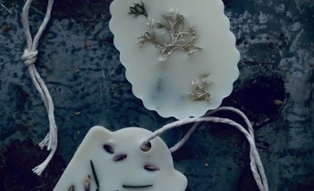 Dwie białe tabliczki z wosku, zakończone zawieszką ze sznurka, z zatopionymi w wosku kawałkami suchych roślin. 
