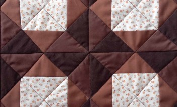 Kawałek brązowej, kwadratowej tkaniny, na której naszyte są cztery małe jasne kwadraty, ułożone w obok siebie w dwóch rzędach. 