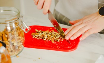 Ręka krojąca nożem orzechy na czerwonej desce. Obok stoi słoik z orzechami.