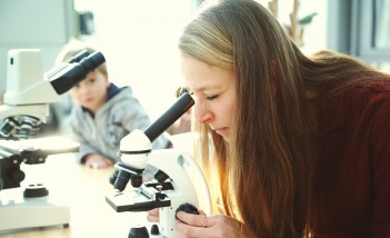 Kobieta patrząca przez mikroskop, za kobietą, na stole, stoi kolejny mikroskop, przy którym siedzi małe dziecko.