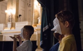 Dwóch chłopców w teatralnych maskach stoi za kurtyną.