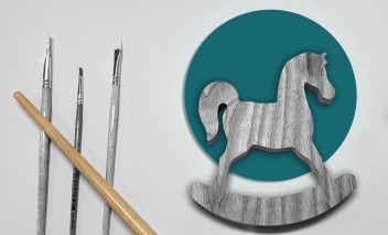 Pośrodku leży drewniany konik, po lewej jest kilka pędzelków, po prawej widać ostrze nożyczek.