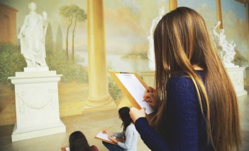 Trzy dziewczyny przed rzeźbą przedstawiającą kobietę owiniętą w szaty szkicują posąg.