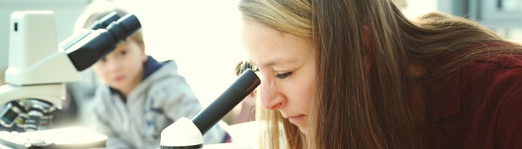 Kobieta patrząca przez mikroskop, za kobietą, na stole, stoi kolejny mikroskop, przy którym siedzi małe dziecko.
